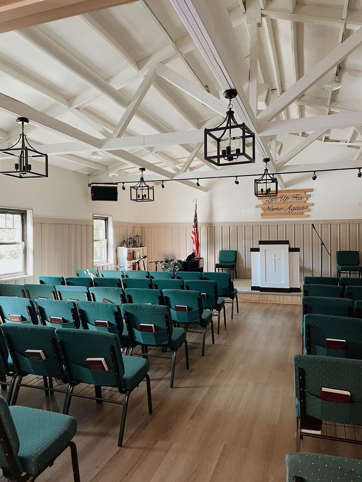 Color photograph of the sanctuary at Landmark Baptist Church in Woodruff: rows of teal padded chairs with Bibles in the seat-back pockets facing a white pulpit with a cross, an American flag and a wooden "Lift Up His Name Again" sign, under white exposed roof trusses and black lantern lights.